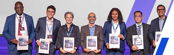 Group of seven professionally dressed individuals standing on stage, each holding a printed report, posing for a formal photo at an event.